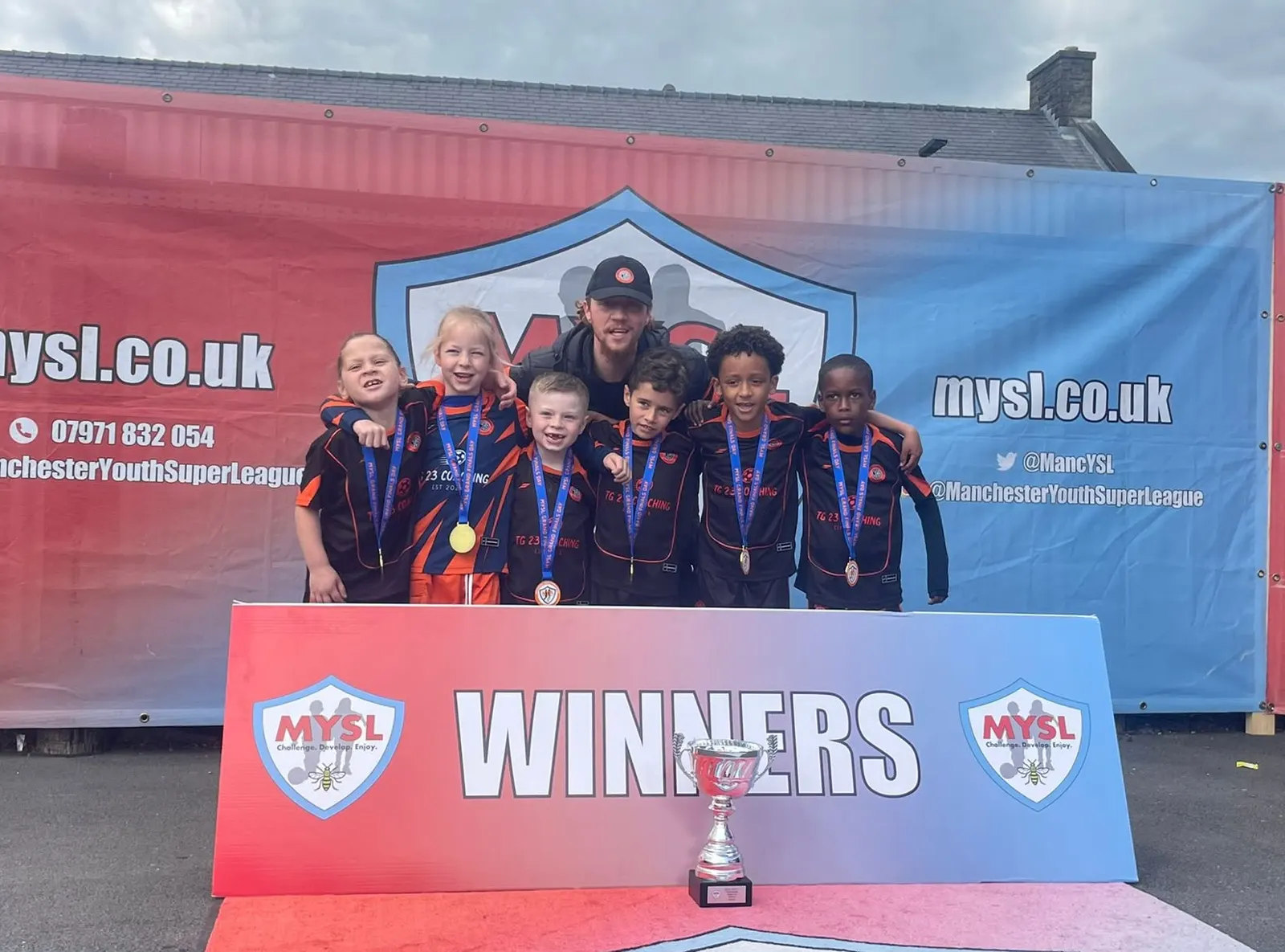 TG23 players and coach with medals and a trophy at a Manchester Youth Super League winners display